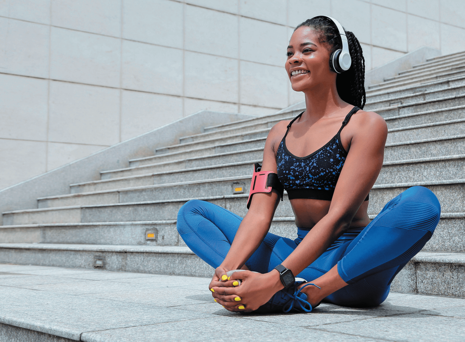 A woman in athletic wear sits cross-legged on outdoor steps, smiling while wearing headphones and stretching.