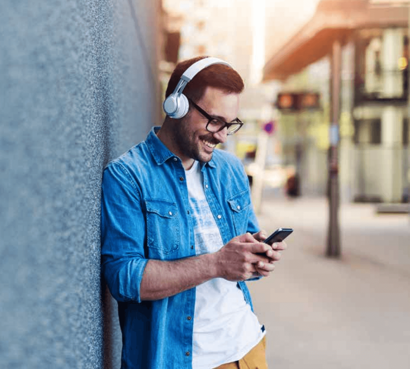 A man wearing headphones leans against a wall outdoors, smiling while looking at his phone.
